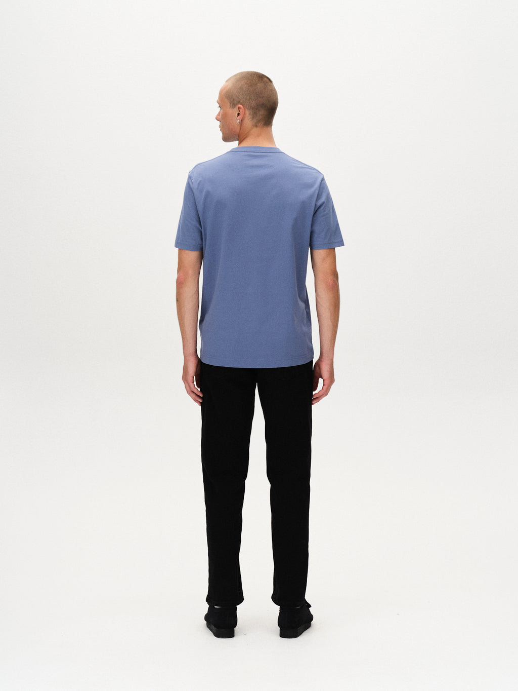 A man stands with his back to the camera against a white background, wearing the Gabba Dune Logo SS GOTS - Blue Horizon t-shirt, black pants, and black shoes.