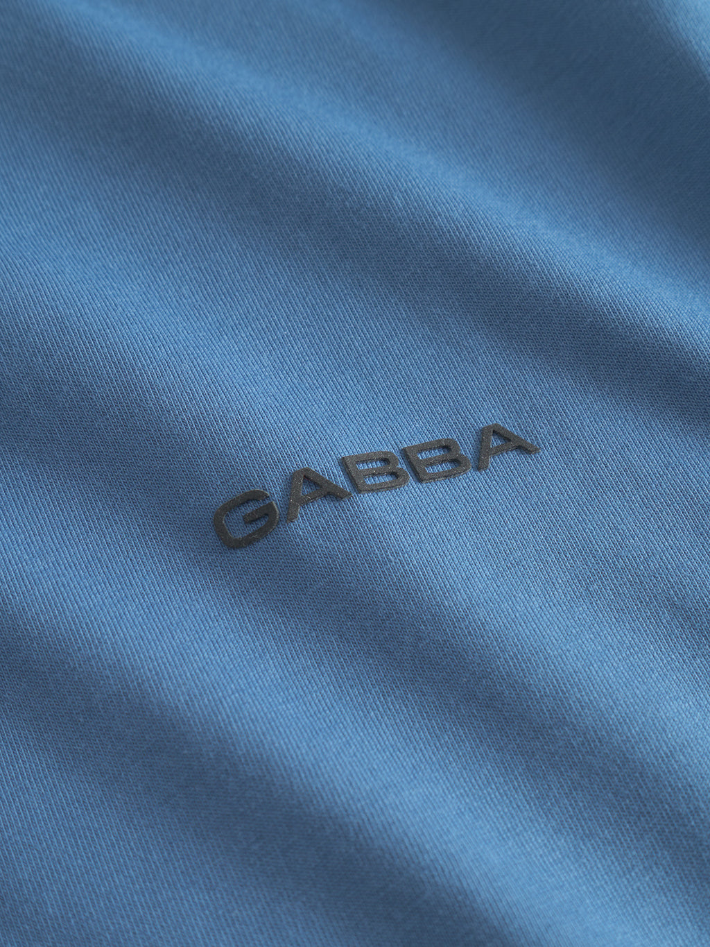 Close-up of Gabbas Dune Logo SS GOTS - Blue Horizon: blue organic cotton fabric with GABBA embroidered in black uppercase letters at the center, evoking a classic t-shirt style.