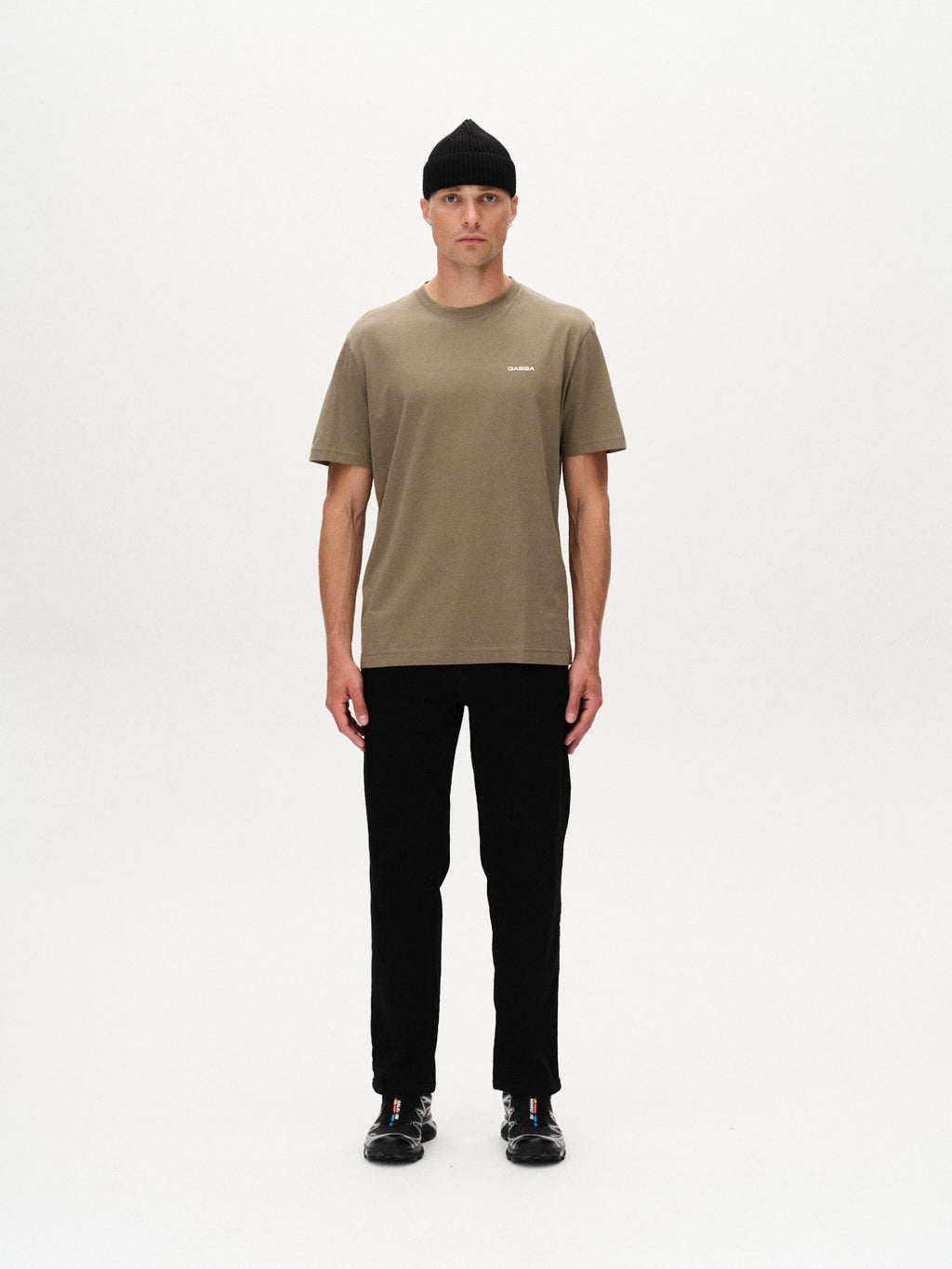 A man stands facing forward against a plain white background, wearing a black beanie, black pants, black sneakers, and a Gabba Dune Logo SS GOTS t-shirt in Deep Lichen Green.