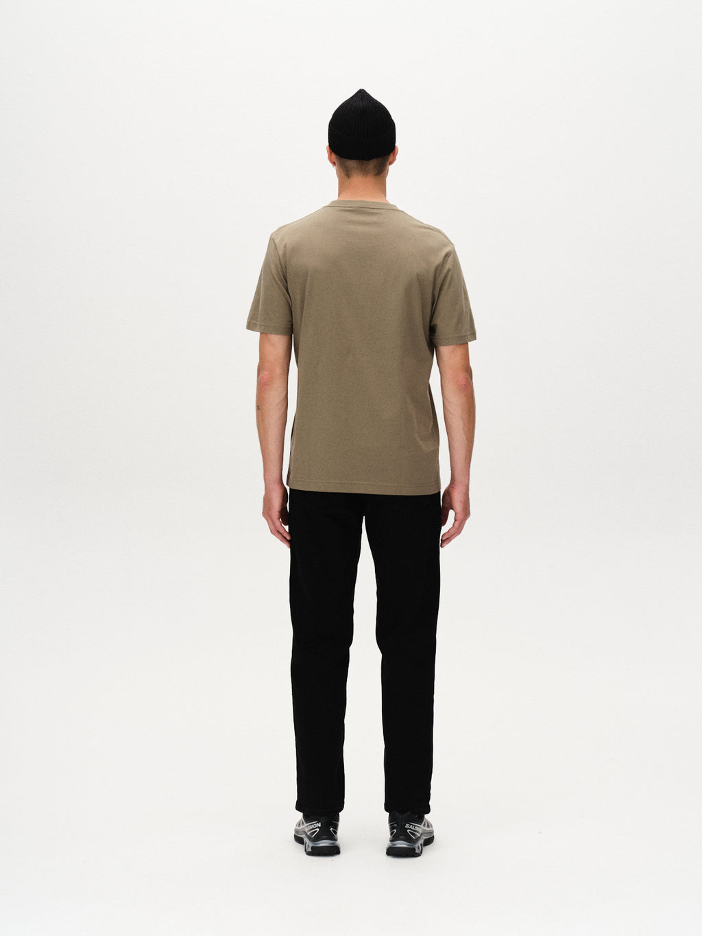 A person wearing a black beanie, the Gabba Dune Logo SS GOTS t-shirt in Deep Lichen Green, black pants, and black sneakers stands facing away from the camera against a plain white background.