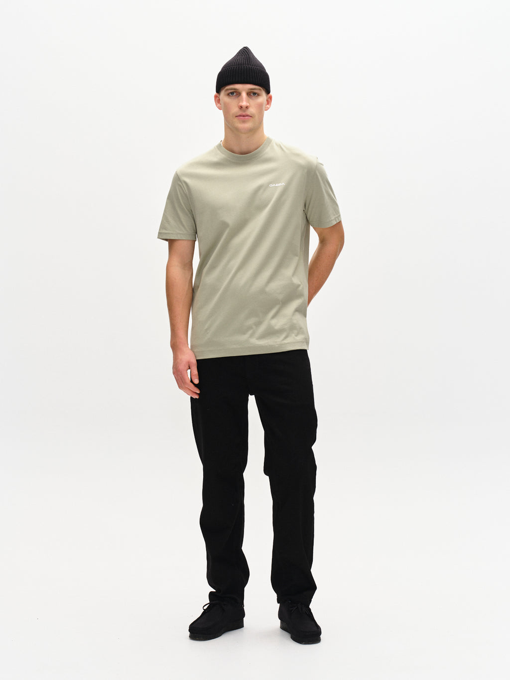 A person stands against a white background, wearing a light green Dune Logo SS GOTS T-shirt by Gabba, with black pants, beanie, and shoes. Their right hand rests by their side while the left is tucked in their pocket.
