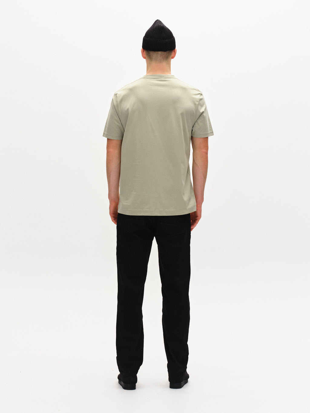 A person wearing the light green Gabba Dune Logo SS GOTS T-shirt, black pants, and a black beanie faces away from the camera against a plain white backdrop.