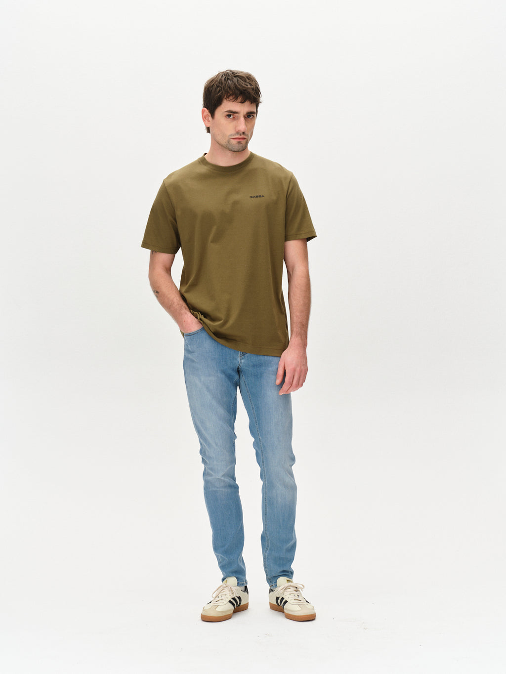 A man stands against a plain white background, wearing Gabbas olive green Dune Logo SS GOTS shirt made of GOTS-certified organic cotton, light blue jeans, and white sneakers with black accents. His left hand is in his pocket while his right arm rests by his side.