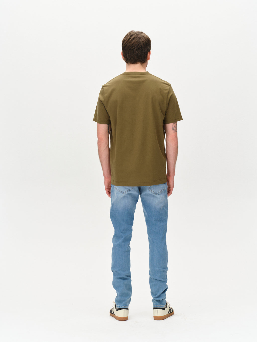 A person with short brown hair, facing away, is wearing Gabbas Dune Logo SS GOTS in Olive Green, paired with light blue jeans and striped sneakers against a plain white backdrop.