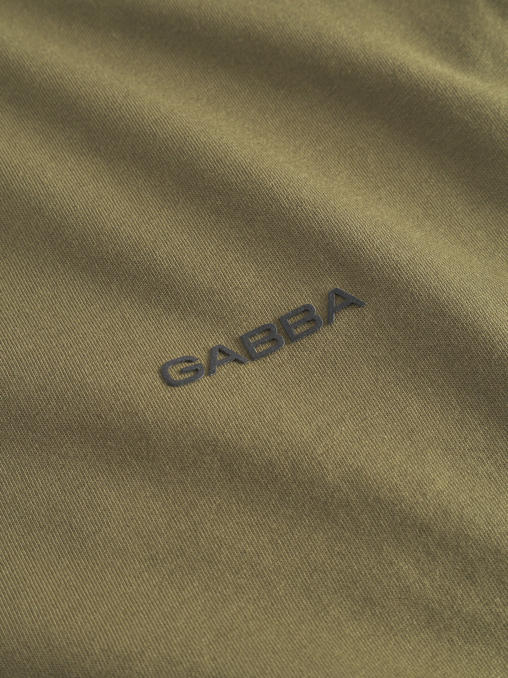 Close-up of the Dune Logo SS GOTS in olive green, by Gabba. Made from GOTS-certified organic cotton, it features the word GABBA embroidered in black thread. The fabric has a smooth texture and slight sheen.