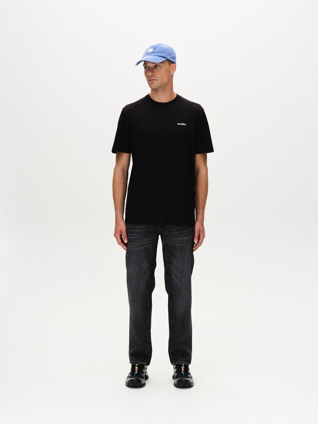 A man stands against a white background, wearing the Gabba Dune Logo SS GOTS black t-shirt. He pairs it with a light blue baseball cap, dark jeans, and black shoes, looking slightly left with his arms relaxed at his sides.