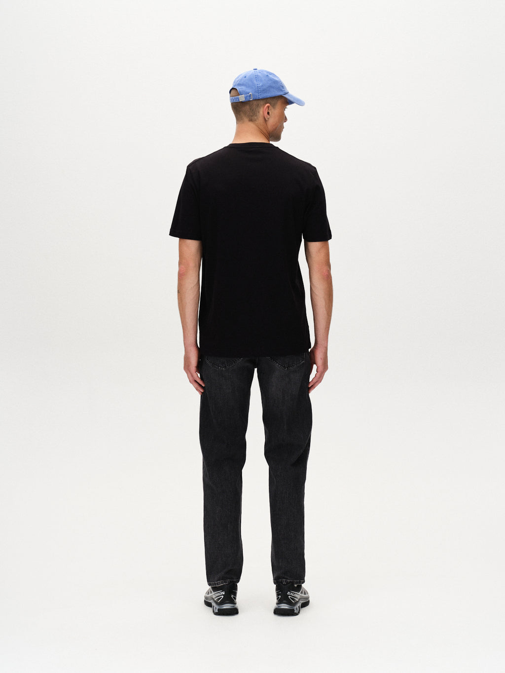 A person stands facing away from the camera, wearing a Gabba Dune Logo SS GOTS - Black classic fit T-shirt, a light blue baseball cap, dark jeans, and black sneakers against a plain white background.