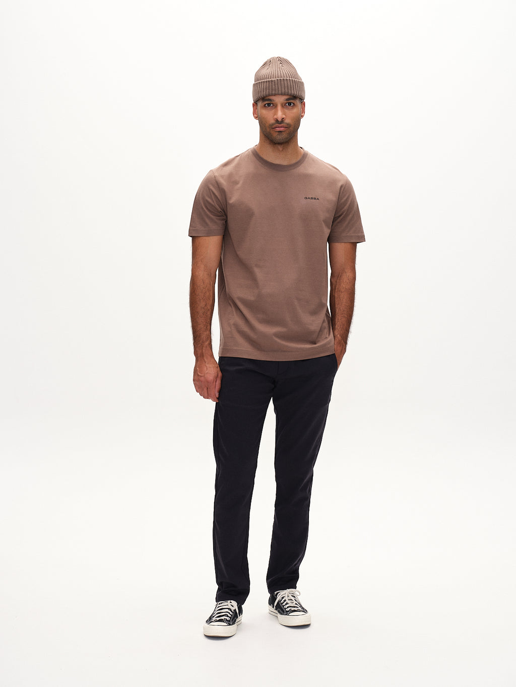 A man stands against a plain background, wearing a Paul Domo - Navy Mel T-shirt by Gabba, slim chino pants with French pockets, a knitted beanie, and sneakers. He looks at the camera with a neutral expression.