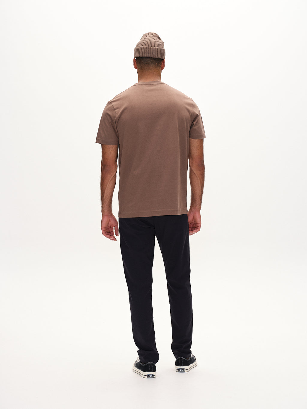 A person stands with their back to the camera, wearing a Gabba Paul Domo - Navy Mel. beige beanie, a brown T-shirt, black slim chino pants with French pockets, and dark sneakers against a plain white background.