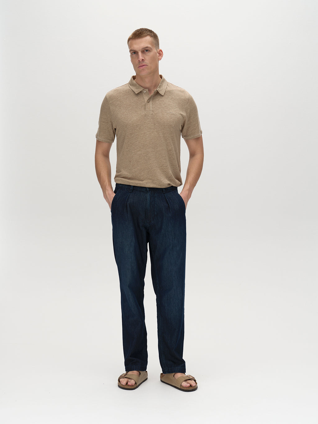 A person with short hair stands against a plain background wearing the Gabba Duke Linen Polo SS in Coriander, dark blue jeans, and sandals. With hands in pockets and a neutral expression, they face straight ahead.