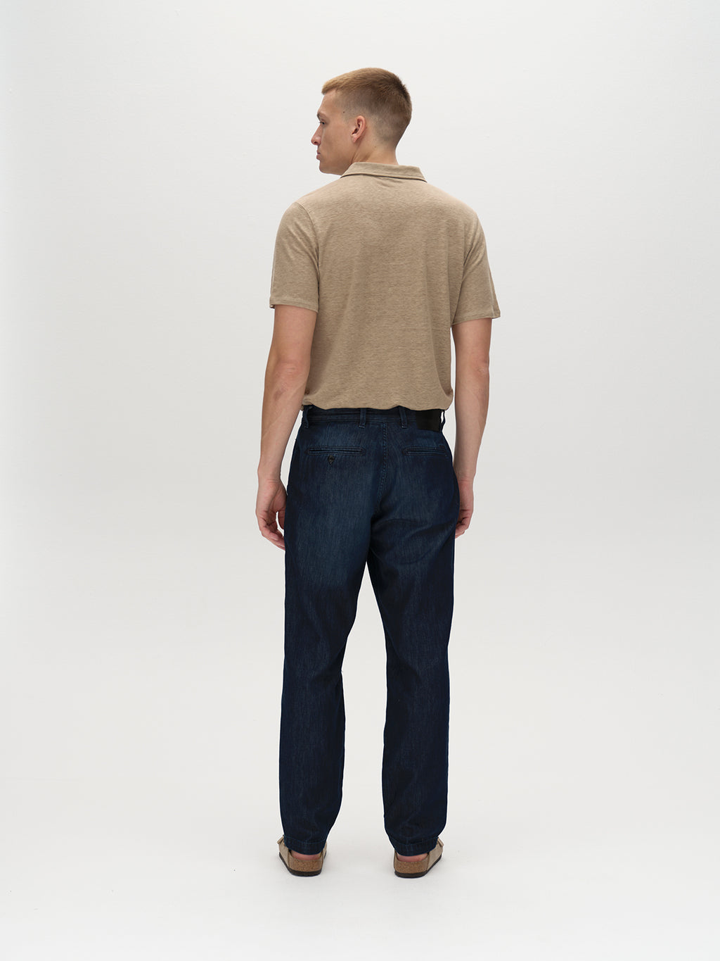A man faces away from the camera, sporting Gabba’s Duke Linen Polo SS in Coriander, complemented by modern fit dark jeans and brown shoes, against a plain white background.