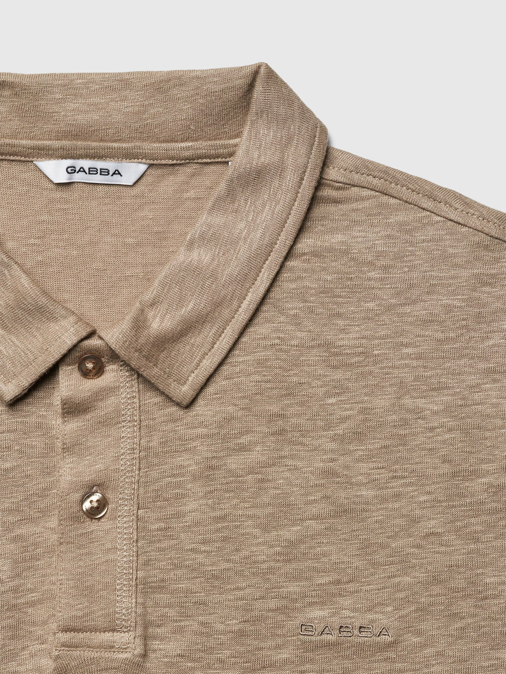 The Duke Linen Polo SS - Coriander by Gabba is a modern fit, beige textured polo with a collared neckline and short sleeves. It features a partially buttoned front with two visible buttons and subtle embroidered branding on the chest, crafted from high-quality fabric.