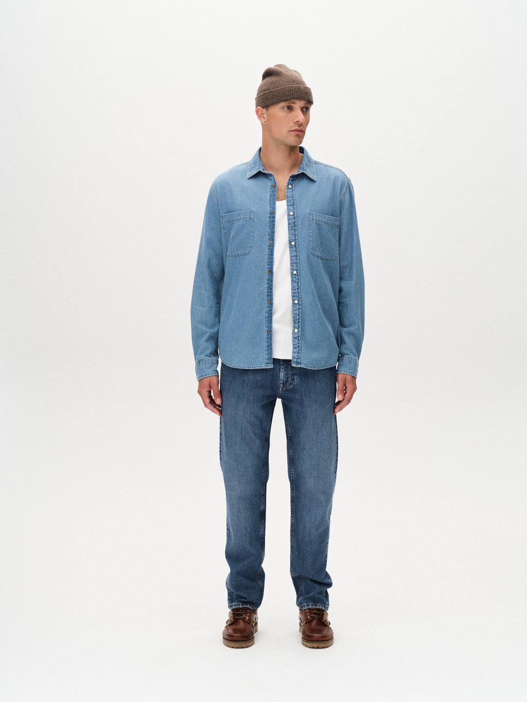 Wearing a brown beanie, white t-shirt, light blue shirt, Marc F1010 - Dark Blue Denim jeans by Gabba, and brown shoes, a person stands against a plain white background for a modern yet classic look.