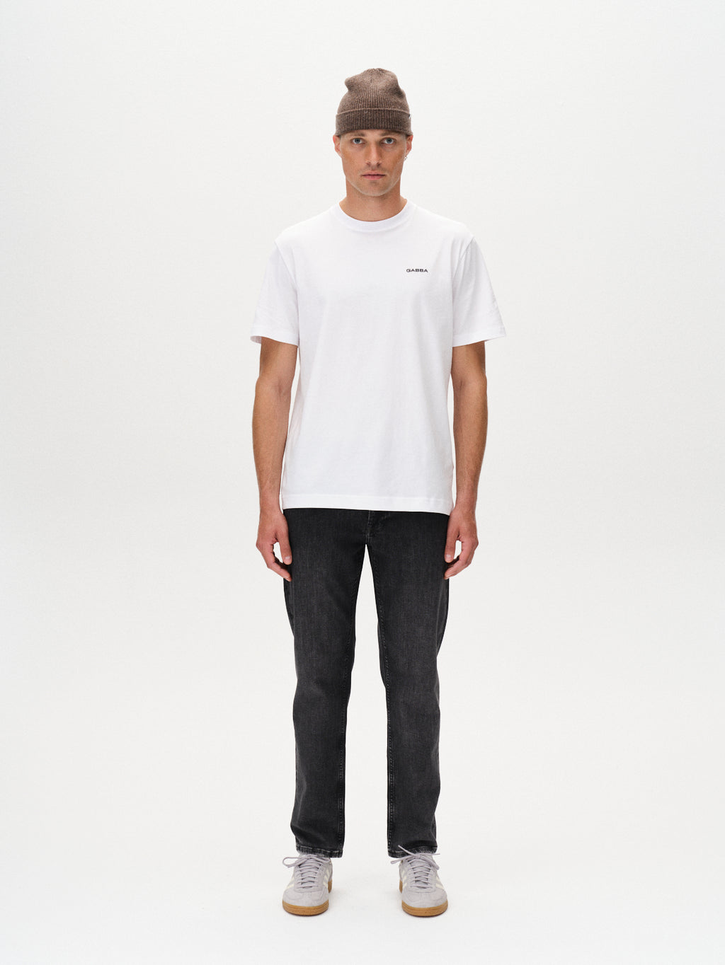 A man stands facing forward against a plain white background, wearing a gray beanie, a white short-sleeve t-shirt, Gabba Marc F1011 - Black Denim jeans, and white sneakers for a modern classic look.