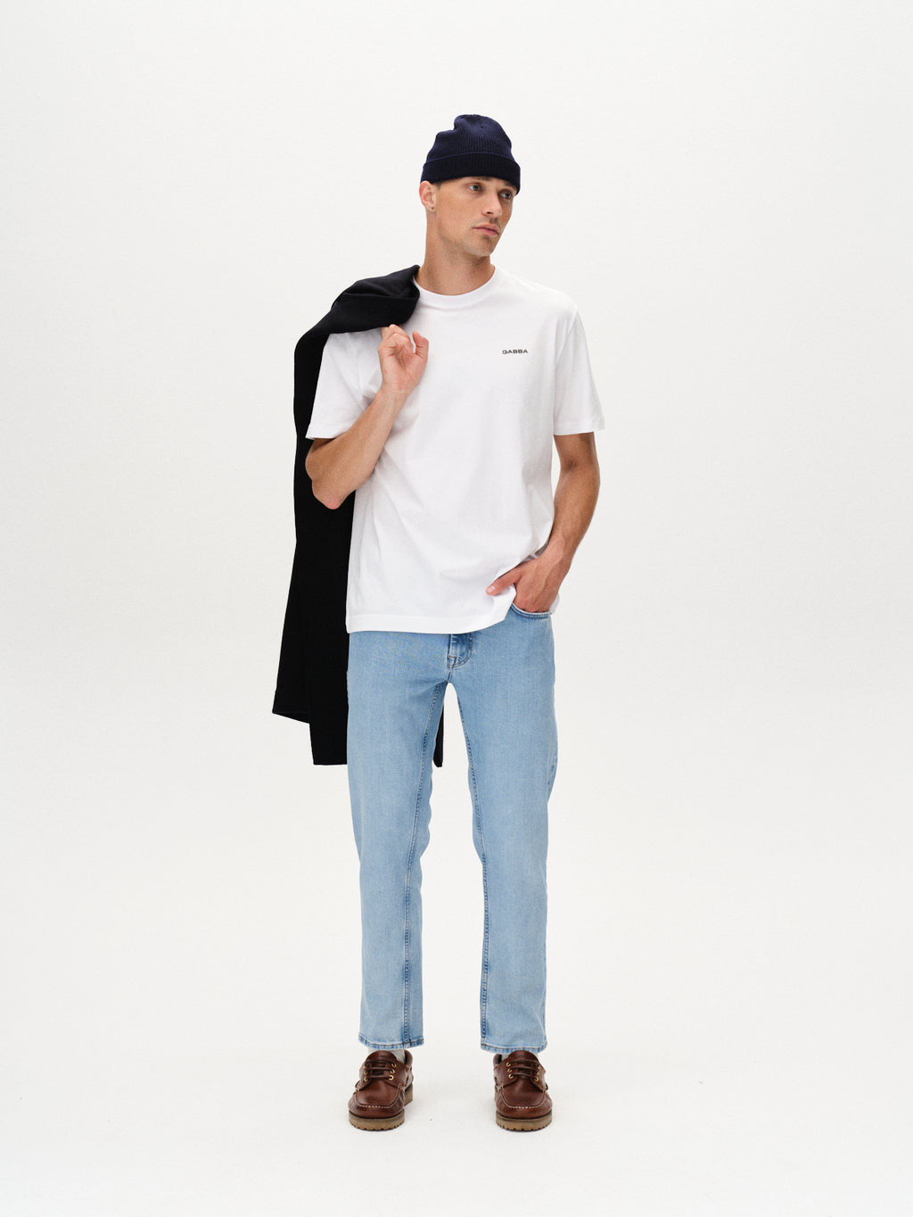 A man stands against a white background, wearing Gabba Marc F1012 - Lt. Blue Denim jeans with a white t-shirt, black beanie, brown shoes, and holding a black jacket over his shoulder for a modern classic look.