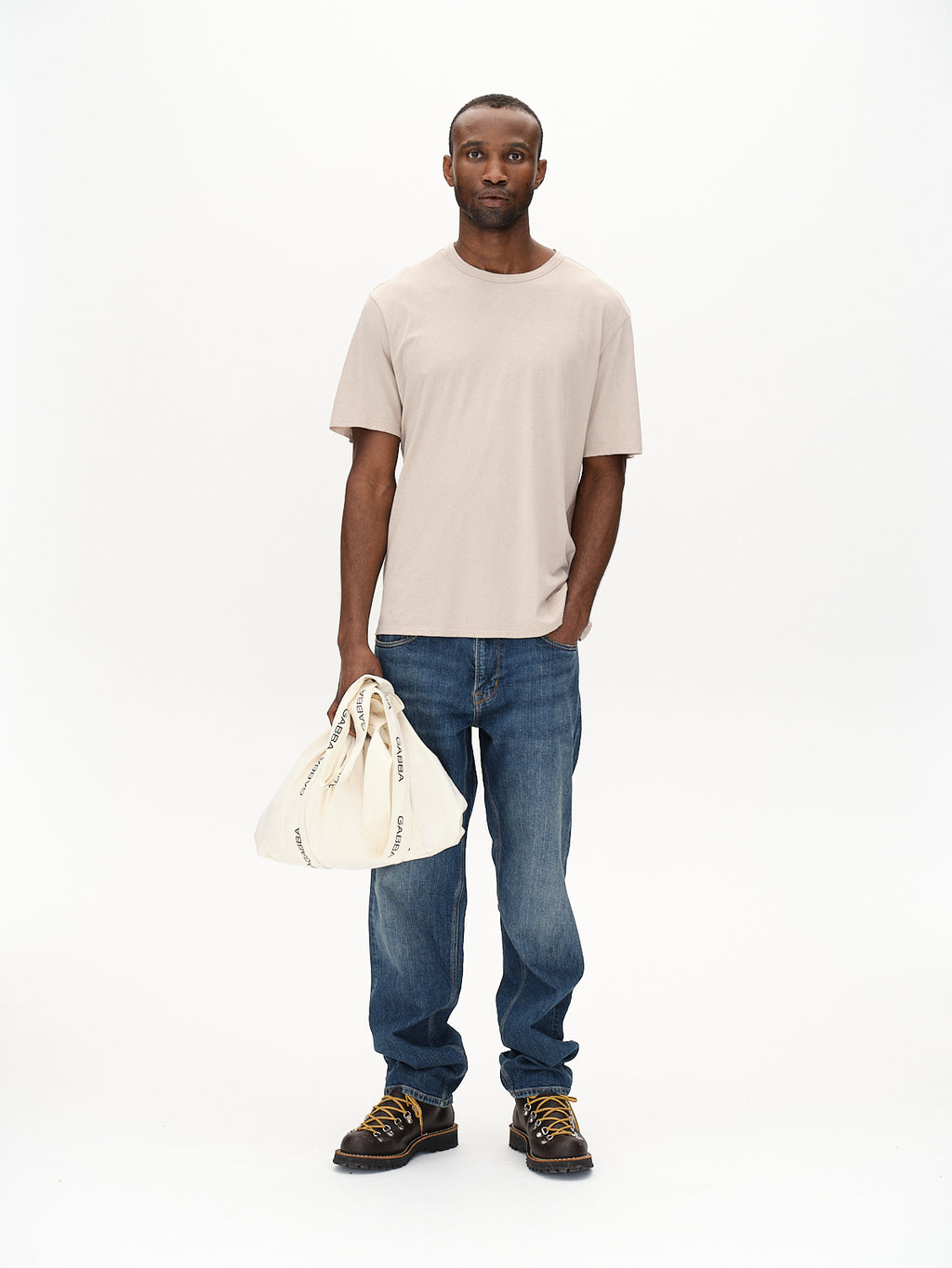 A man stands against a white background, wearing Gabbas Lagos SS - Sand T-shirt and blue jeans, showcasing versatile style. Brown hiking boots promise comfort, while he holds a beige cloth bag in his right hand. His expression is neutral.