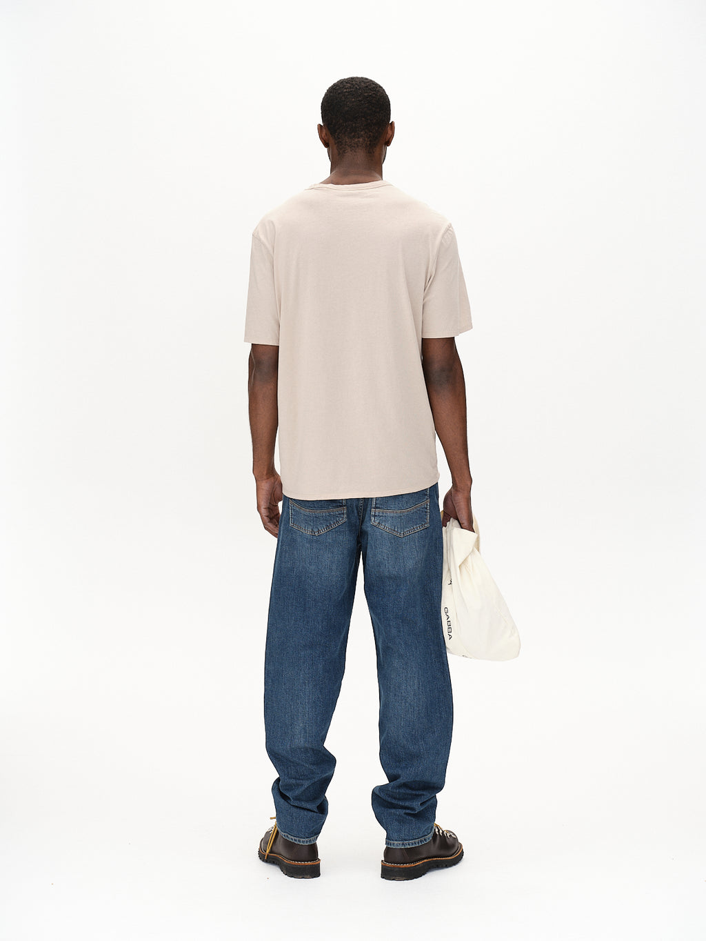 Facing away from the camera, a person wears the Gabba Lagos SS - Sand t-shirt paired with loose blue jeans for all-day comfort. They hold a white tote bag in their left hand, showcasing a versatile wardrobe style against a plain white background.
