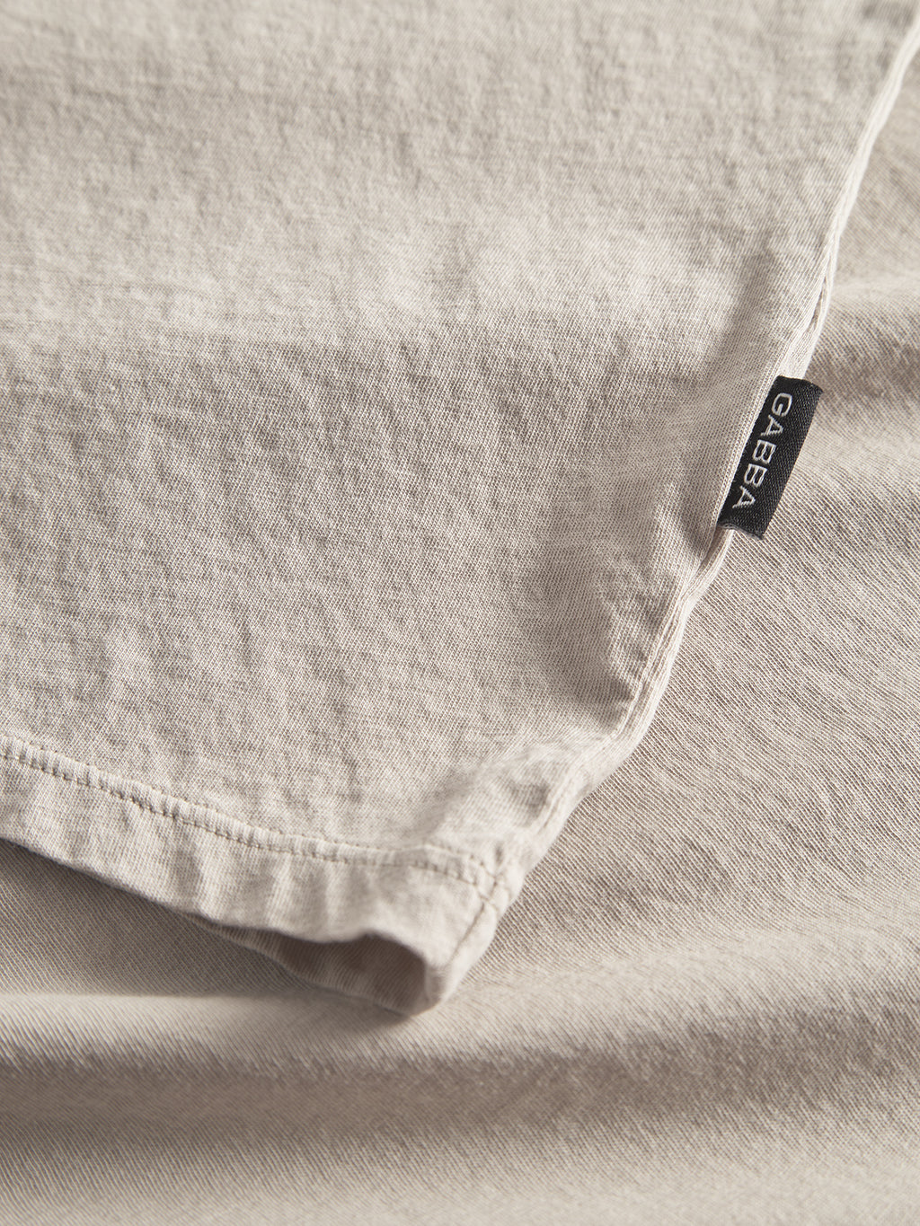 Close-up of Lagos SS - Sand by Gabba, a light beige fabric with subtle texture. This classic fit material offers a stitched edge and features a small black tag with GABBA in white, ensuring all-day comfort for your versatile wardrobe.