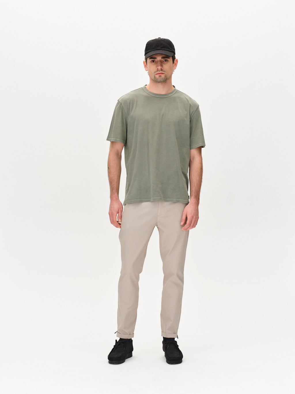 A man wears the Gabba Lagos SS - Green Sea, a classic fit T-shirt with a comfortable round neckline, paired with beige pants, black cap, and black shoes, standing against a plain white background.