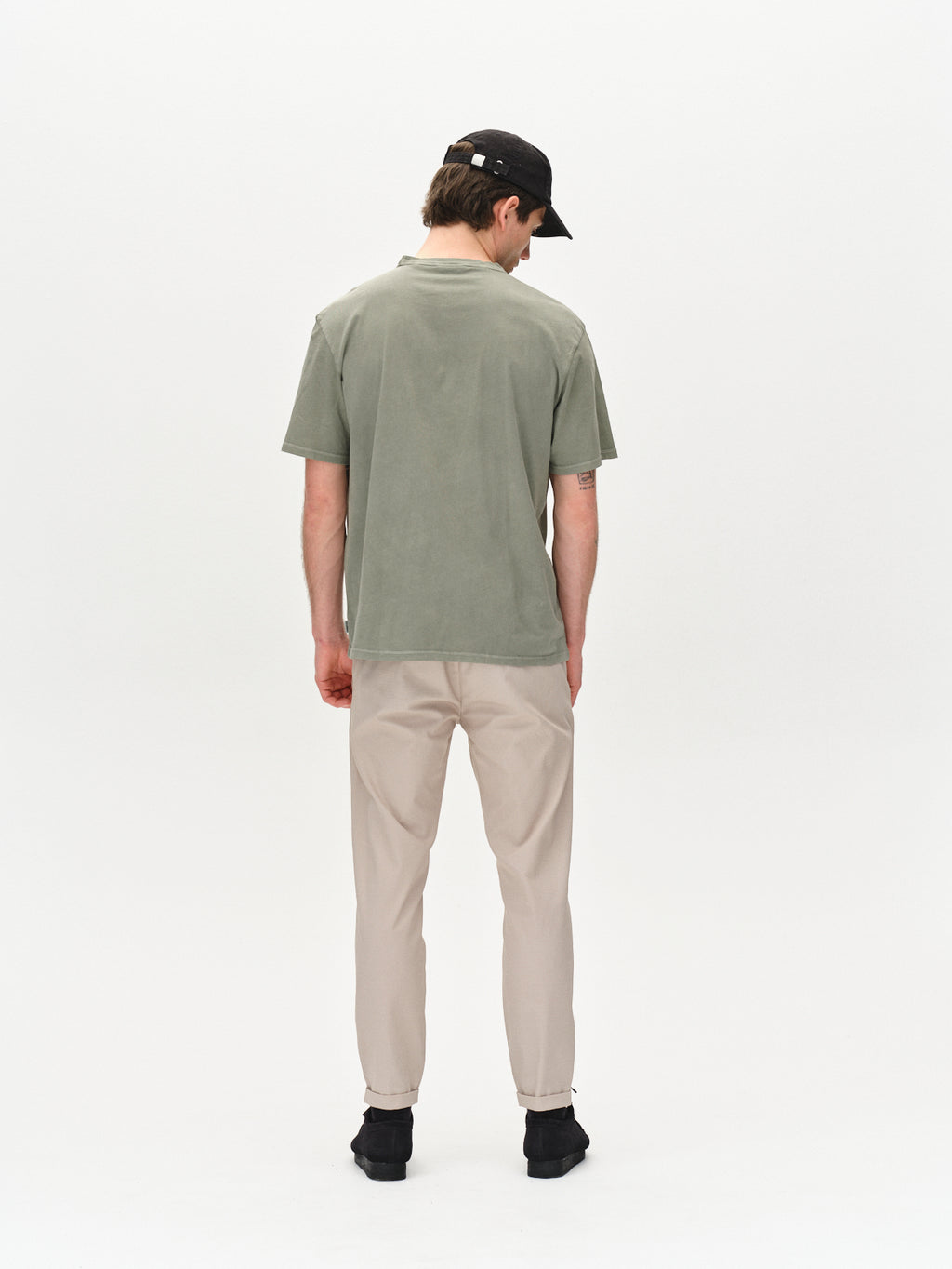A man in a classic fit Lagos SS - Green Sea T-shirt by Gabba with a comfortable round neckline, beige pants, black cap, and shoes stands facing away against a plain white background.