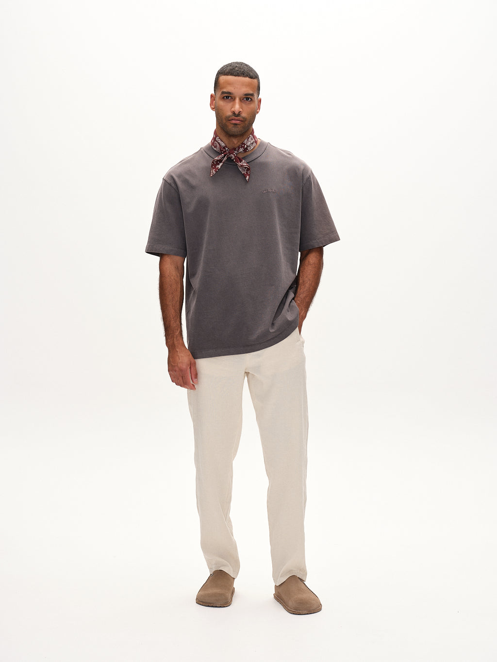 Against a plain white background, a man exudes classic comfort in his signature style. He wears a gray T-shirt with Gabbas Napoli 19 Twill Linen pants in Birch, brown shoes, and a patterned bandana around his neck, with his hands resting by his sides.