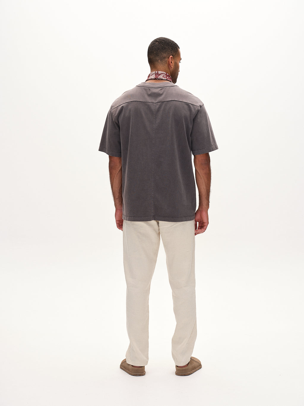 A man showcases a classic style wearing a gray short-sleeve shirt, Napoli 19 Twill Linen - Birch pants by Gabba, and brown shoes. A red patterned scarf adds flair to his neck. The plain white background highlights the ensembles comfort and elegance.