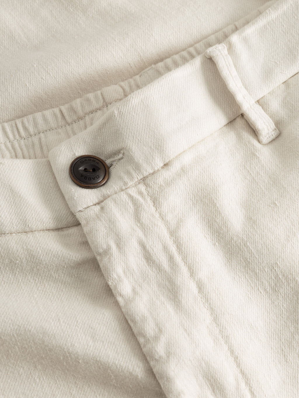 A close-up of Gabbas Napoli 19 Twill Linen pants in Birch highlights the waistband with a four-hole brown button and fabric belt loop. The textured twill suggests durable comfort, combining classic style with linen craftsmanship.