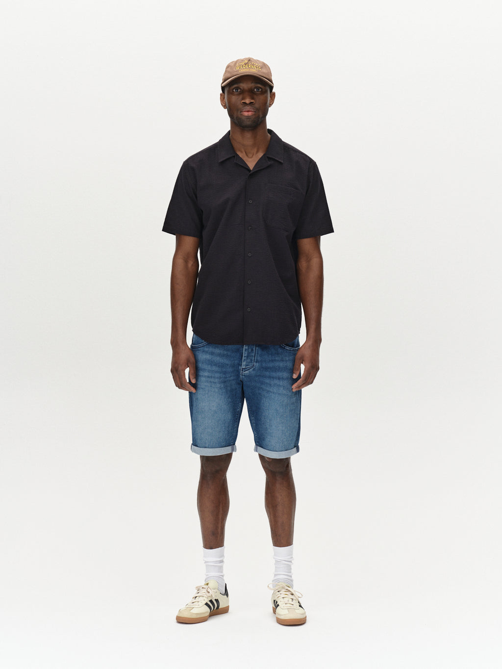A person stands wearing a black short-sleeve shirt, Gabbas Jason K3787 SANZA Shorts in Mid Blue Denim, white socks, beige sneakers, and a beige cap. The plain background emphasizes the casual spring and summer ensemble.