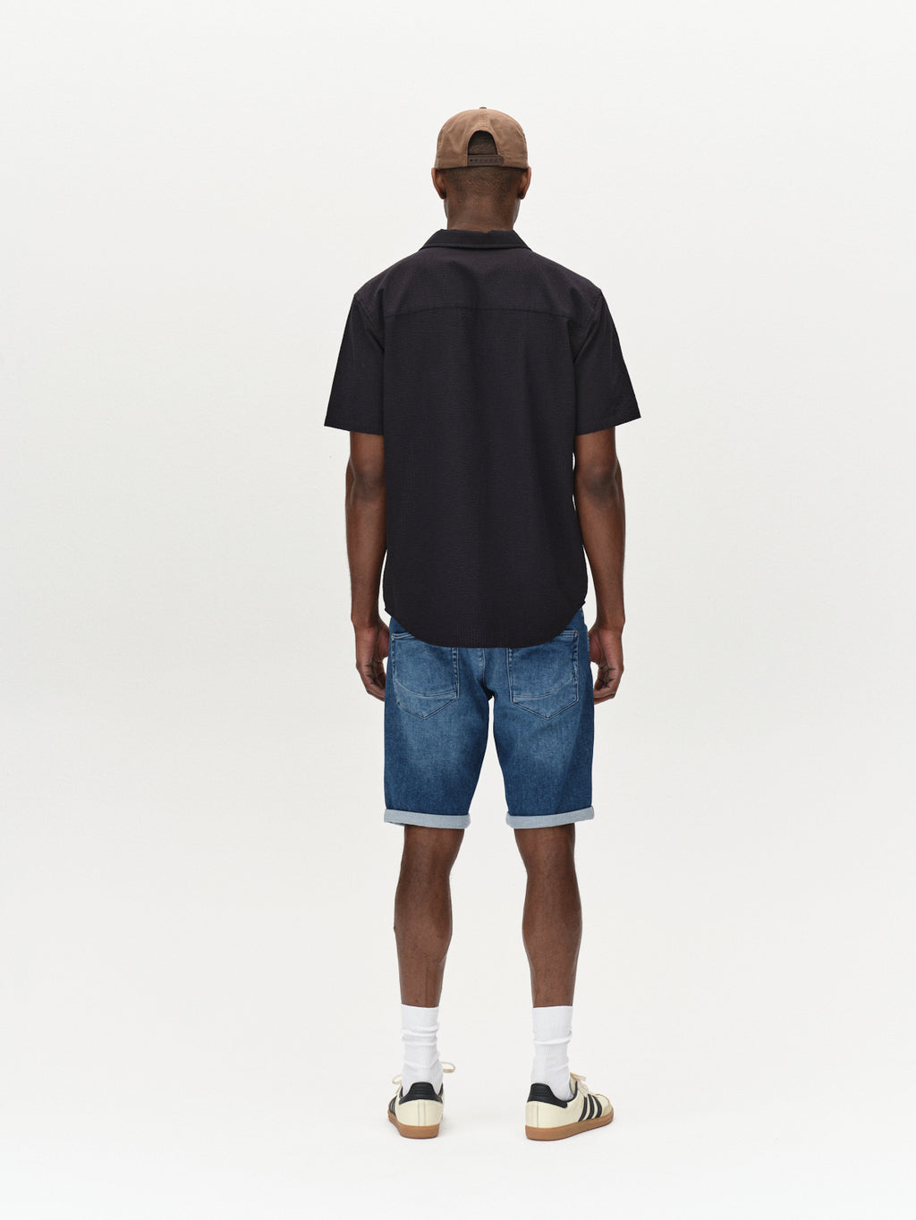 Someone, wearing a cap, a Gabba black short-sleeve shirt, and Jason K3787 SANZA Shorts in Mid Blue Denim by Gabba stands facing away against a plain white background. Beige sneakers and white socks complete their perfect spring and summer look.