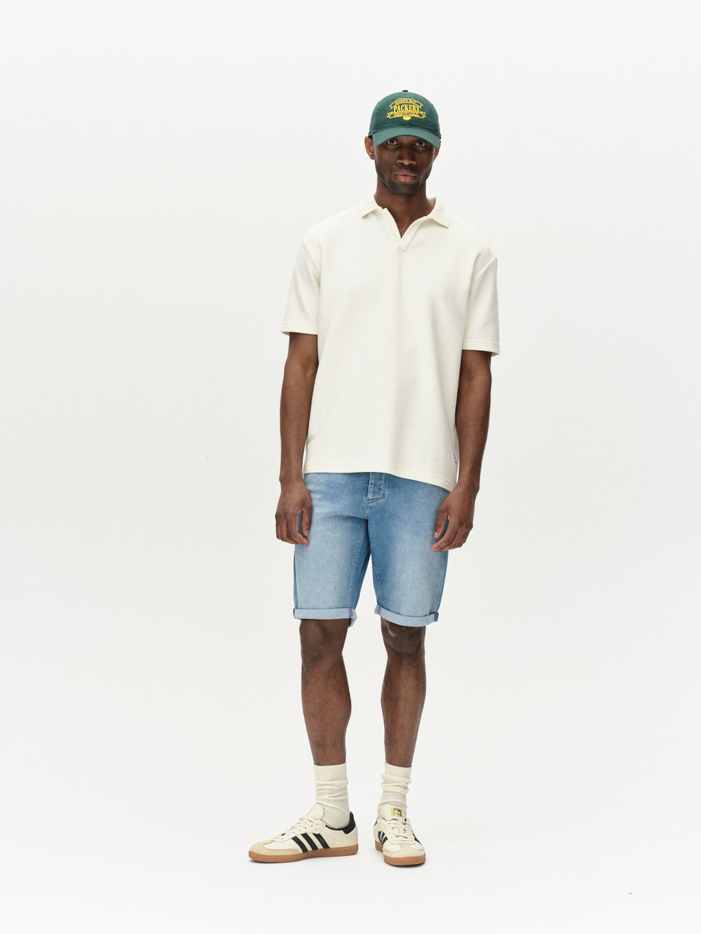 A man stands against a plain white background, wearing a green cap, white short-sleeve polo shirt, Gabba Jason K3787 SANZA Shorts in light blue denim, white socks, and beige sneakers with black stripes.