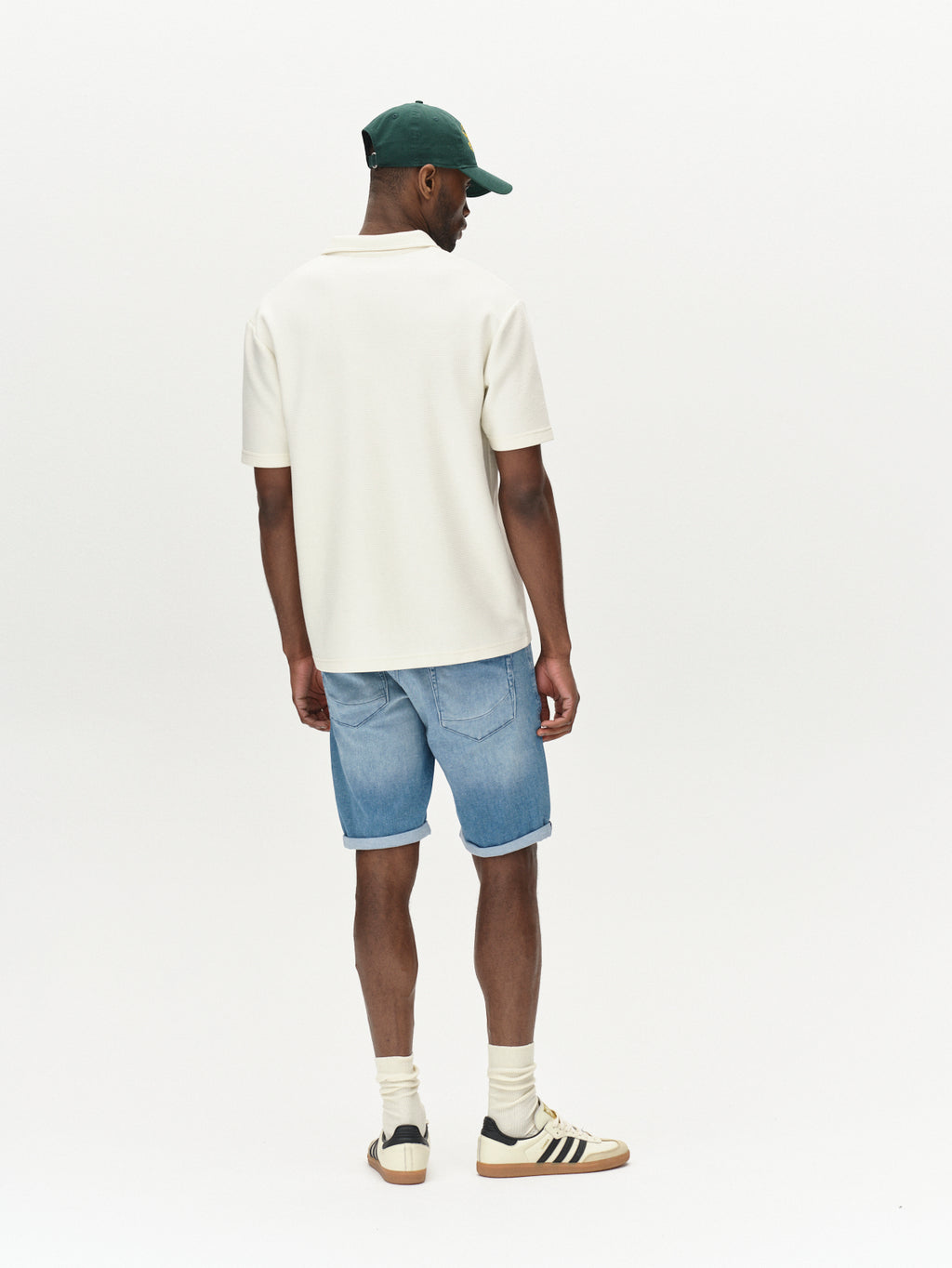 A man in a white short-sleeve shirt, Gabba Jason K3787 SANZA Shorts in light blue denim, white socks, sneakers, and a green cap stands facing away against a plain light background.