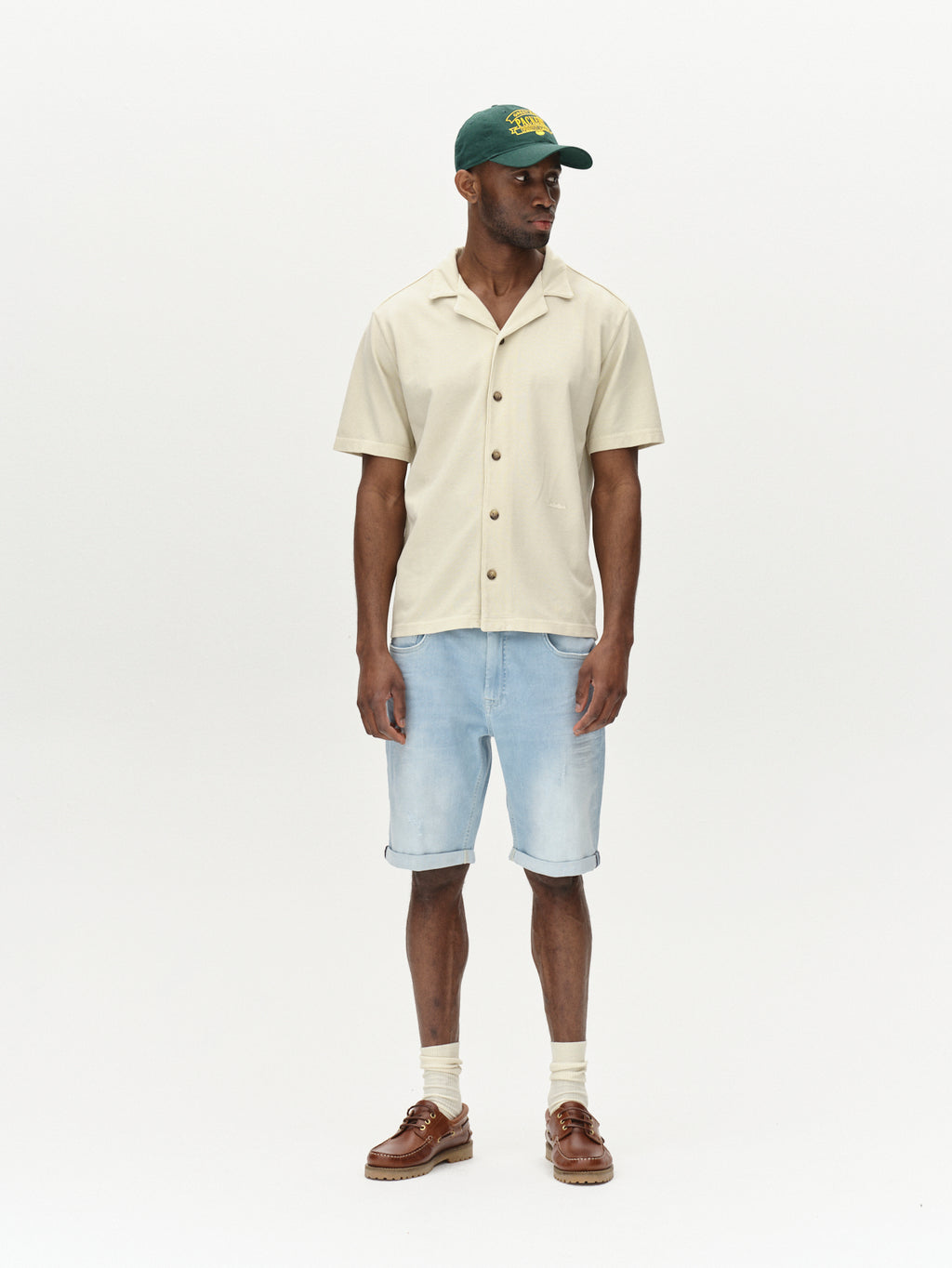 A man in a beige short-sleeve button-up shirt and Gabbas Markus K3987 Shorts in light blue denim stands against a plain background. Completing his look with a green cap, brown shoes, and light-colored socks, his hands rest relaxed by his sides, perfect for any laid-back day.