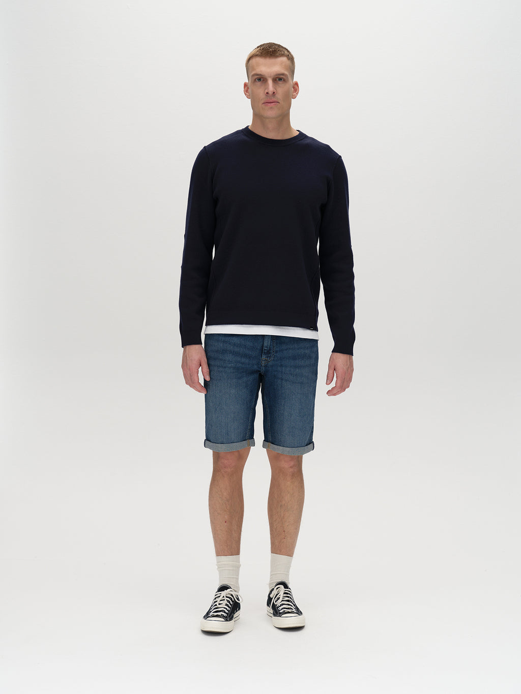 A person stands against a plain background, wearing a navy sweater over a white shirt, Gabbas Markus K4664 Shorts in Mid Blue Denim, and black sneakers with white socks. They face forward with a neutral expression.