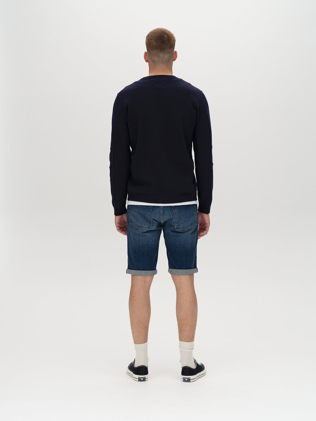A person stands facing away, wearing a cozy dark sweater, Markus K4664 Shorts in mid blue denim by Gabba, white socks, and black sneakers against a plain white background.