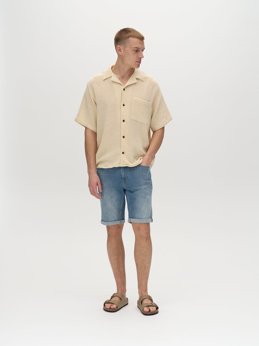 A person stands against a white background wearing a beige short-sleeve button-up shirt, Gabbas Markus K4664 Shorts in light blue denim, and brown sandals. They have short hair and are gazing slightly to the side.