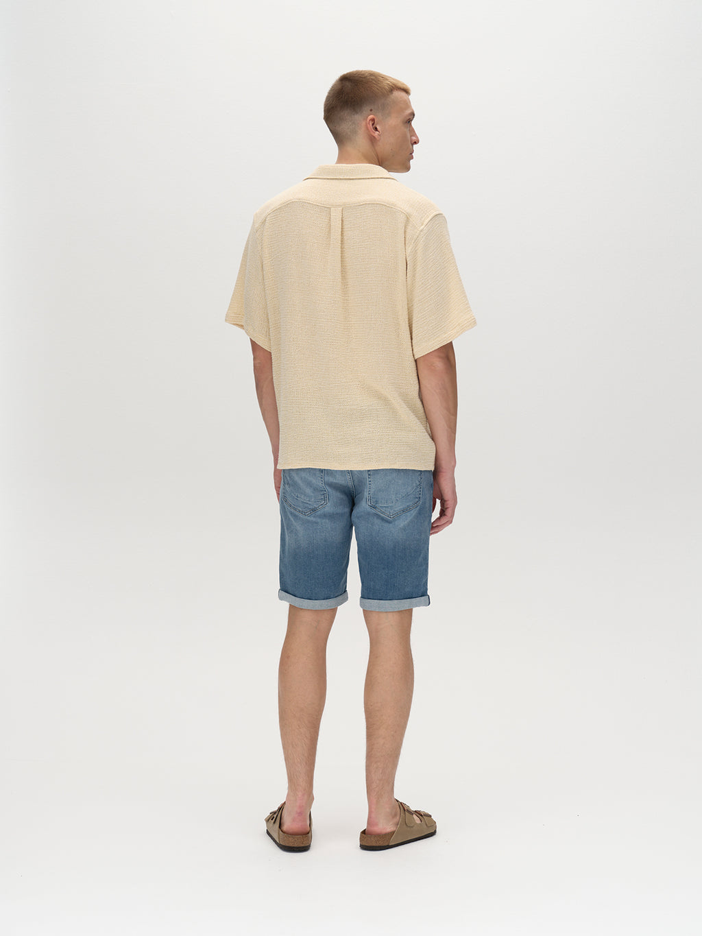 A man, showcasing Gabbas Markus K4664 Shorts in light blue denim, wears a beige short-sleeve shirt and brown sandals. The ensemble highlights a comfortable summer look against a plain, light-colored background.