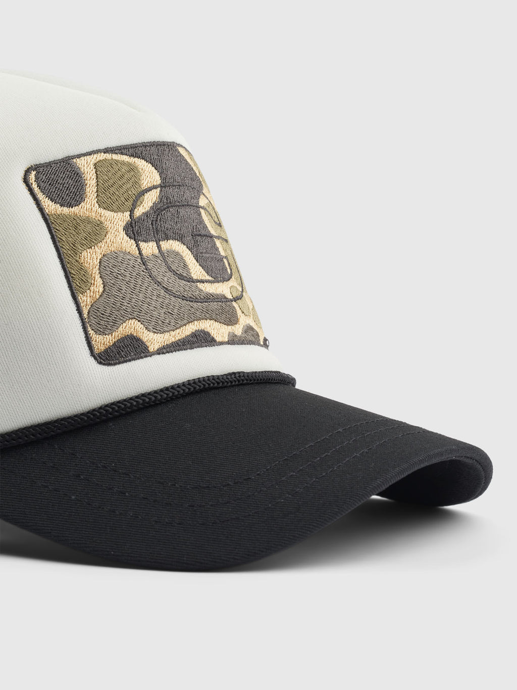 A close-up of the Wolf Cam - Meteorite baseball cap shows a black visor and light crown, featuring an embroidered circular camouflage patch with the Gabba logo. Its adjustable strap ensures a perfect fit for any head size.