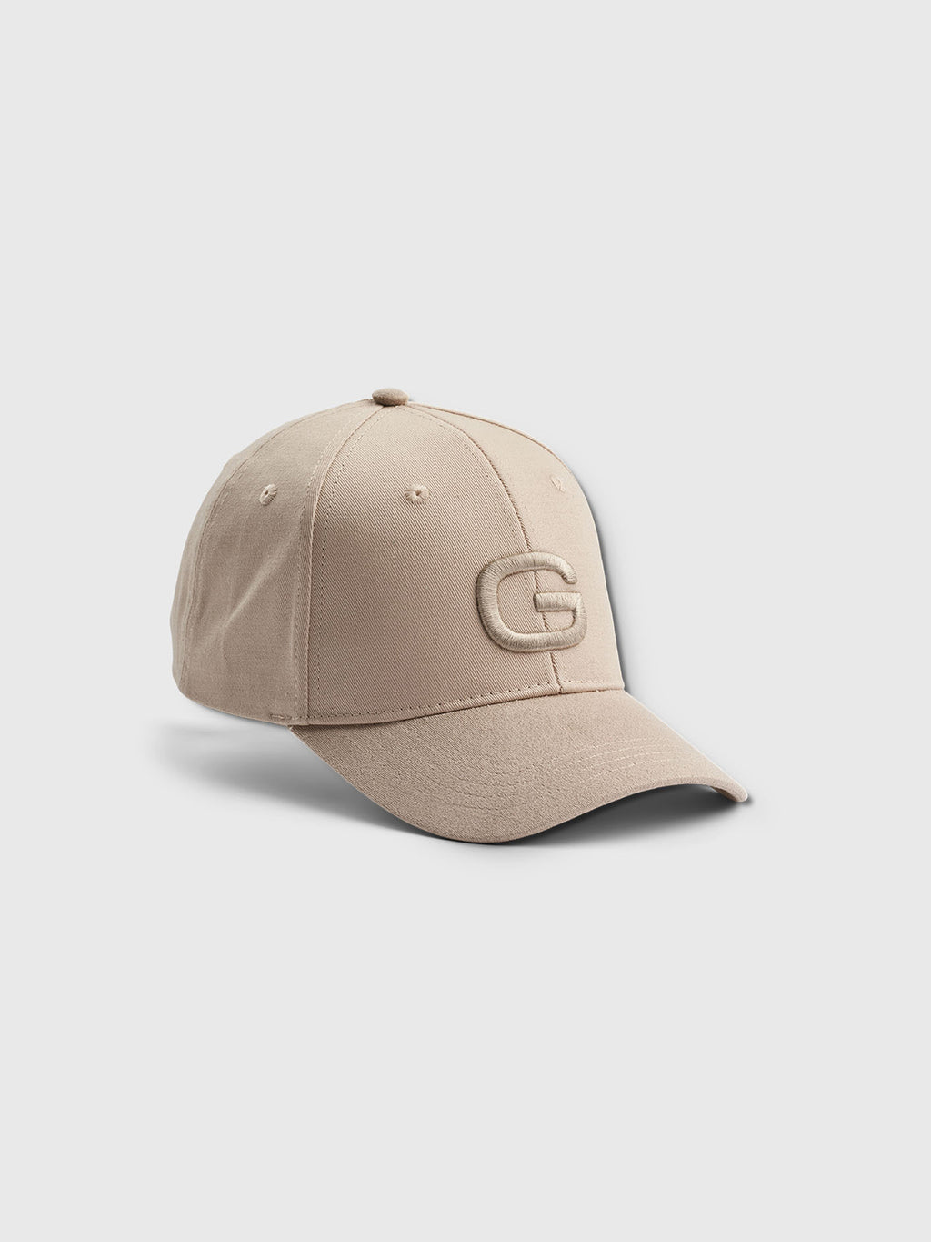 The Gabba Base - Sand cap boasts a classic fit with a curved brim and an embroidered G in beige. Its modern design is highlighted as it rests at an angle on a plain white background.