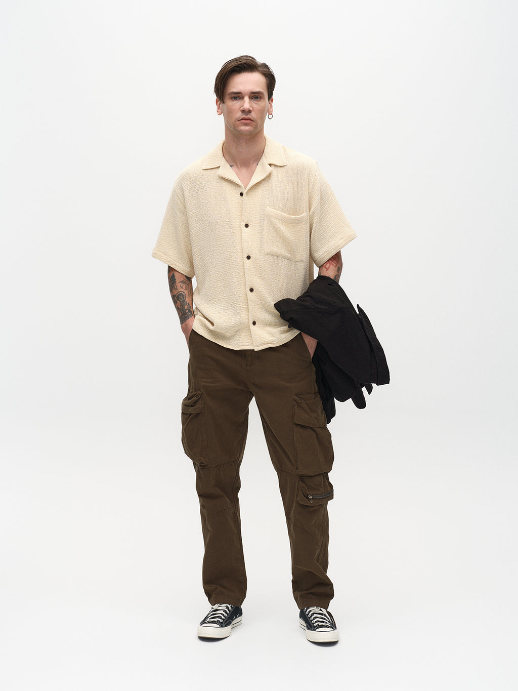 Against a plain backdrop, a person sports a beige short-sleeve button-up shirt, Gabbas Rodi Cargo Pant in Grape Leaf with functional pockets, and black sneakers. Holding a dark jacket, their outfit of durable cotton fabric highlights their tattoos and short hair.