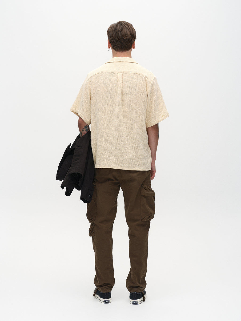 A person with short hair, in a beige short-sleeved shirt and Gabbas Rodi Cargo Pant - Grape Leaf, is seen from the back. They hold a black jacket in their right hand against a plain white background.