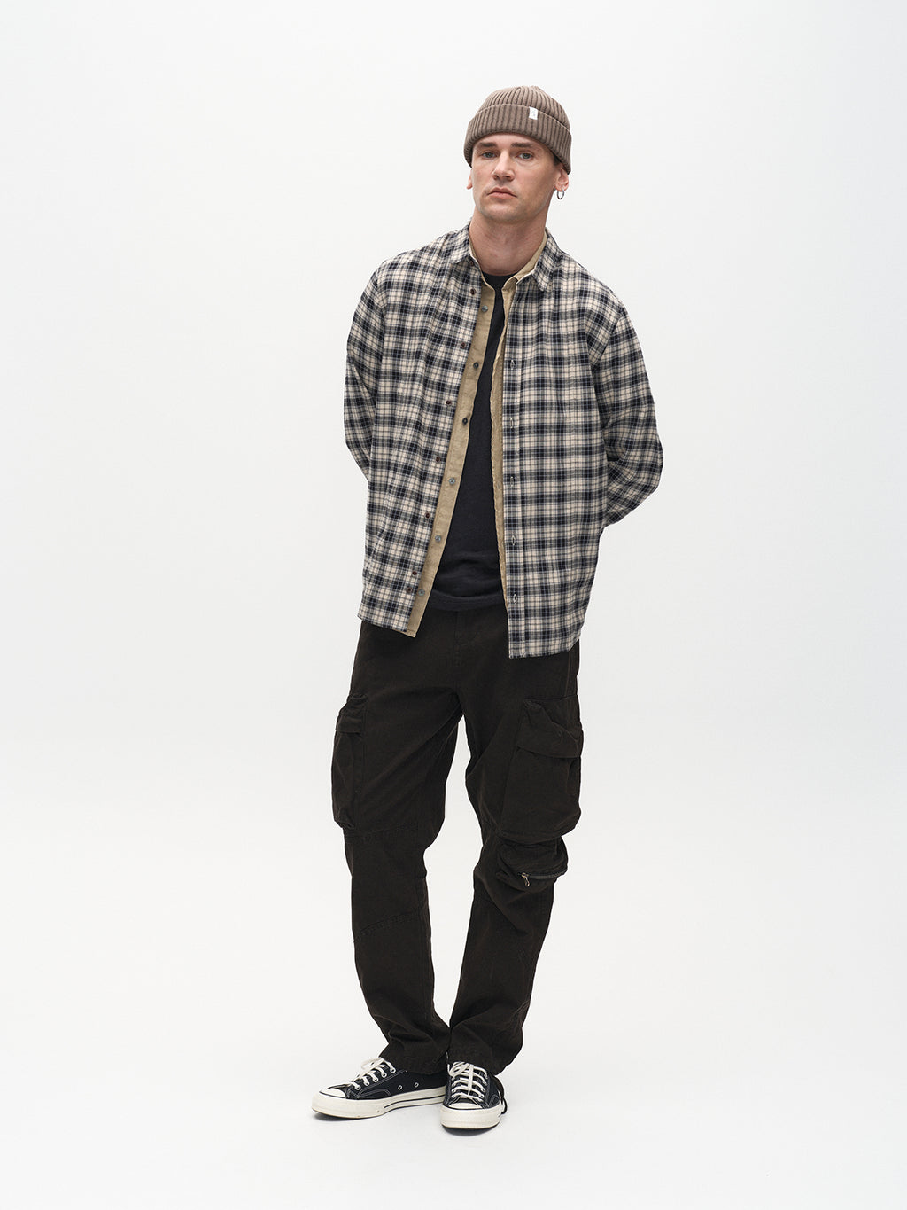 A man stands against a white background, wearing a plaid button-up shirt over a black tee, Gabbas Rodi Cargo Pant in black comfort stretch cotton, black Converse sneakers, and a brown beanie. His hands are behind his back as he gazes slightly to the side.