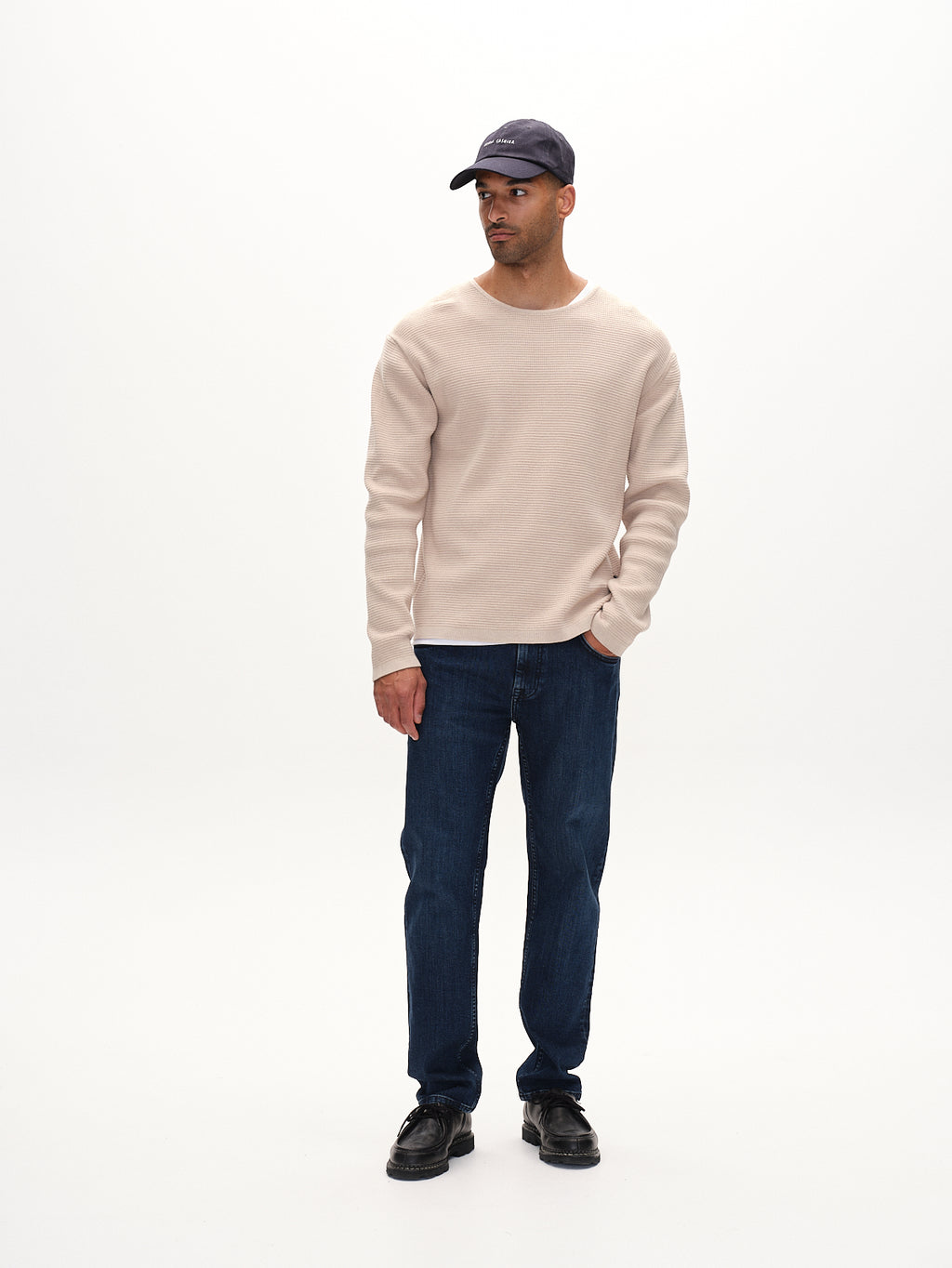 A man in a Gabba Lambo LS - Birch classic jumper pairs it with dark blue jeans, black shoes, and a dark baseball cap. Standing against a plain white background and looking to his left, he embodies a seamless blend of contemporary flair and timeless charm.