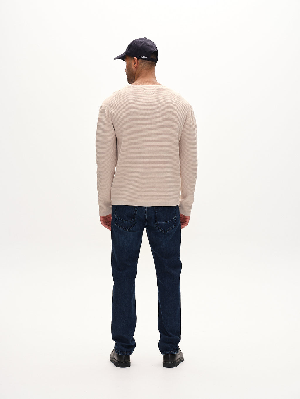 A man faces away from the camera against a plain white background, dressed in Gabbas Lambo LS - Birch jumper crafted with contemporary techniques, paired with dark blue jeans, black shoes, and a dark cap.