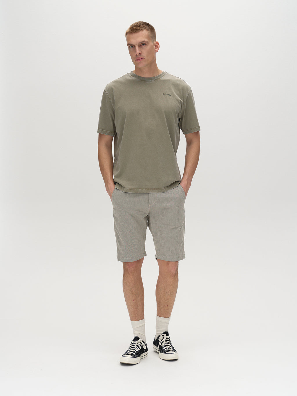 A person with short hair stands against a plain background, embodying modern design in an olive green t-shirt and Gabbas Jet K3280 Dale Shorts in Birch. The comfortable fit is completed with white socks and black sneakers, hands casually tucked into pockets while looking forward.