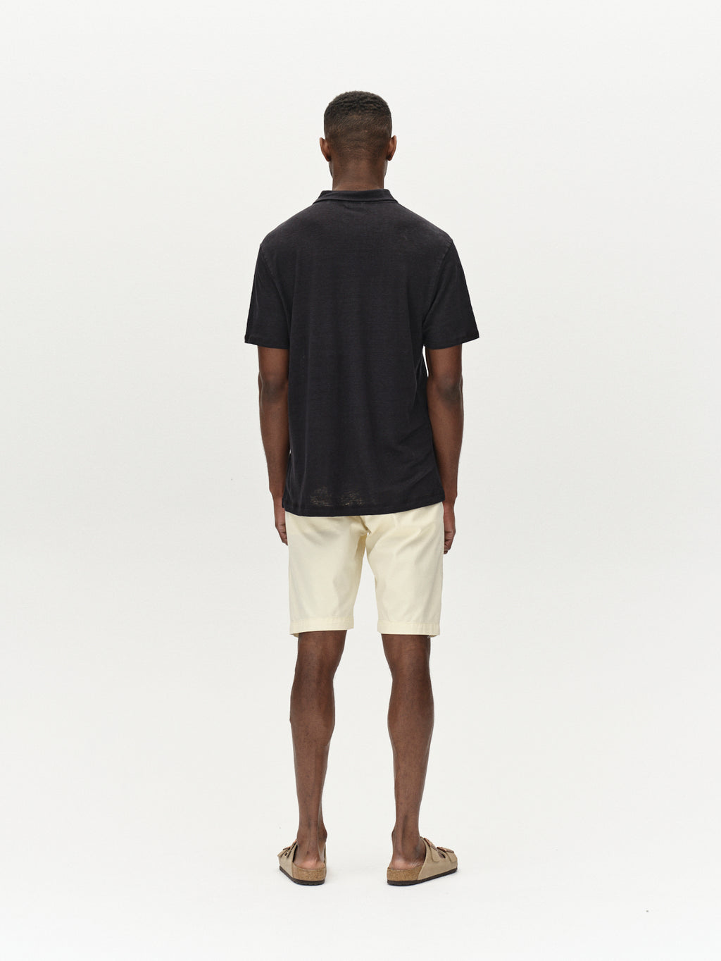 A man stands facing away against a white background, wearing a black short-sleeve shirt, Jet K3280 Dale Shorts - Ecru Light from Gabba, and brown sandals. The comfortable fit exemplifies versatile design, perfect for any casual occasion.