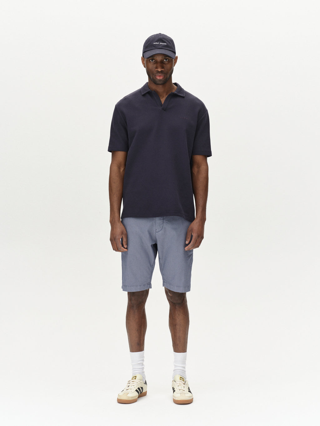 A man poses against a plain backdrop, wearing a dark short-sleeve polo with Gabbas Jet K3280 Dale Shorts in blue, epitomizing timeless appeal. His ensemble is rounded off with white socks, beige sneakers, and a dark baseball cap. Hands at his sides and a neutral expression exude comfort.