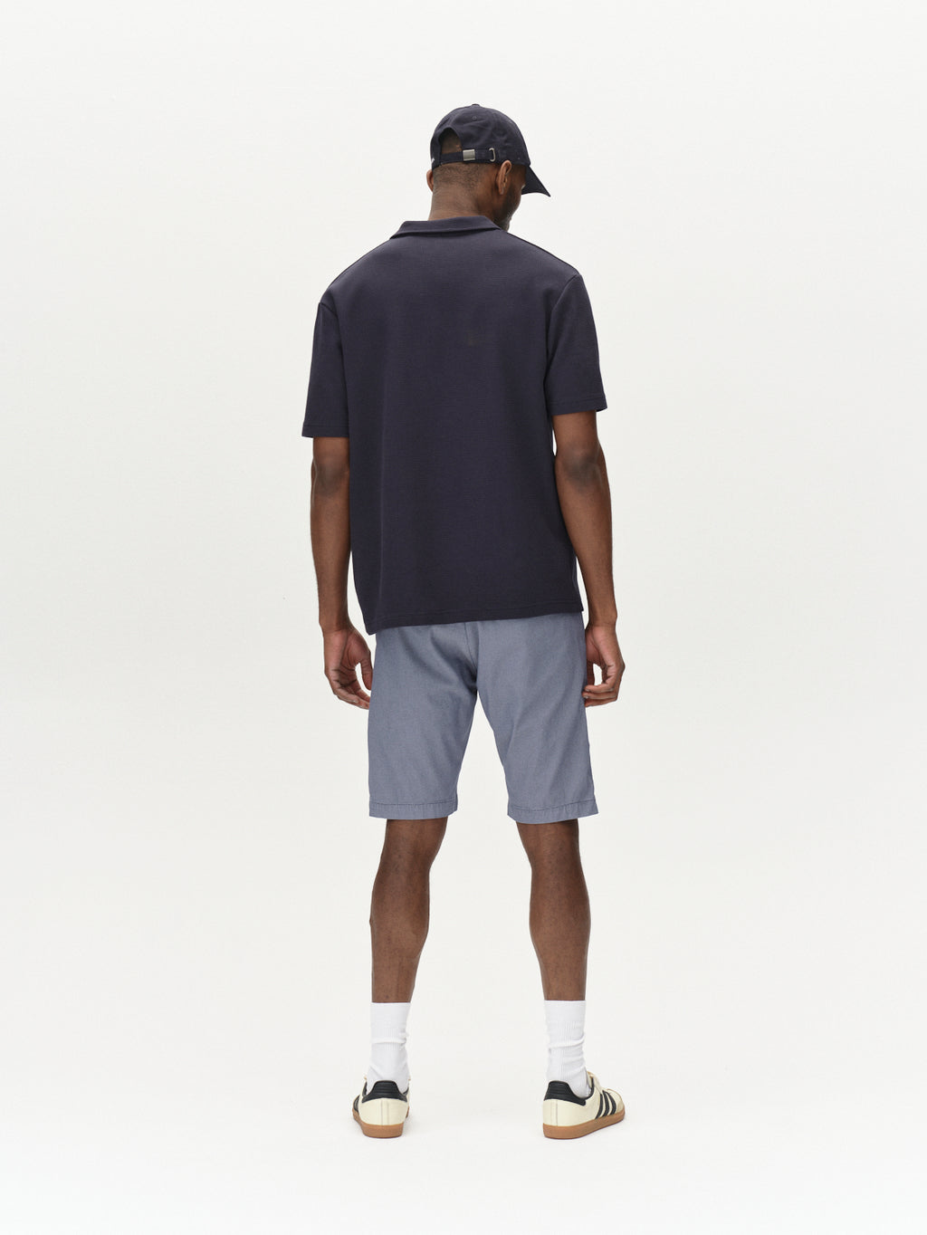 A person, back to the camera, sports a black cap, dark polo, and Gabbas Jet K3280 Dale Shorts - Blue for a comfy fit. White socks and sneakers complete this timeless look against a plain, light background.
