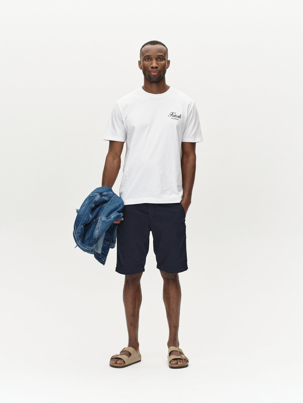 A man stands against a plain white background in a T-shirt with text on the chest and Gabbas Jet K3280 Dale Shorts in navy for all-day comfort. He completes his modern look with beige sandals and holds a blue jacket.