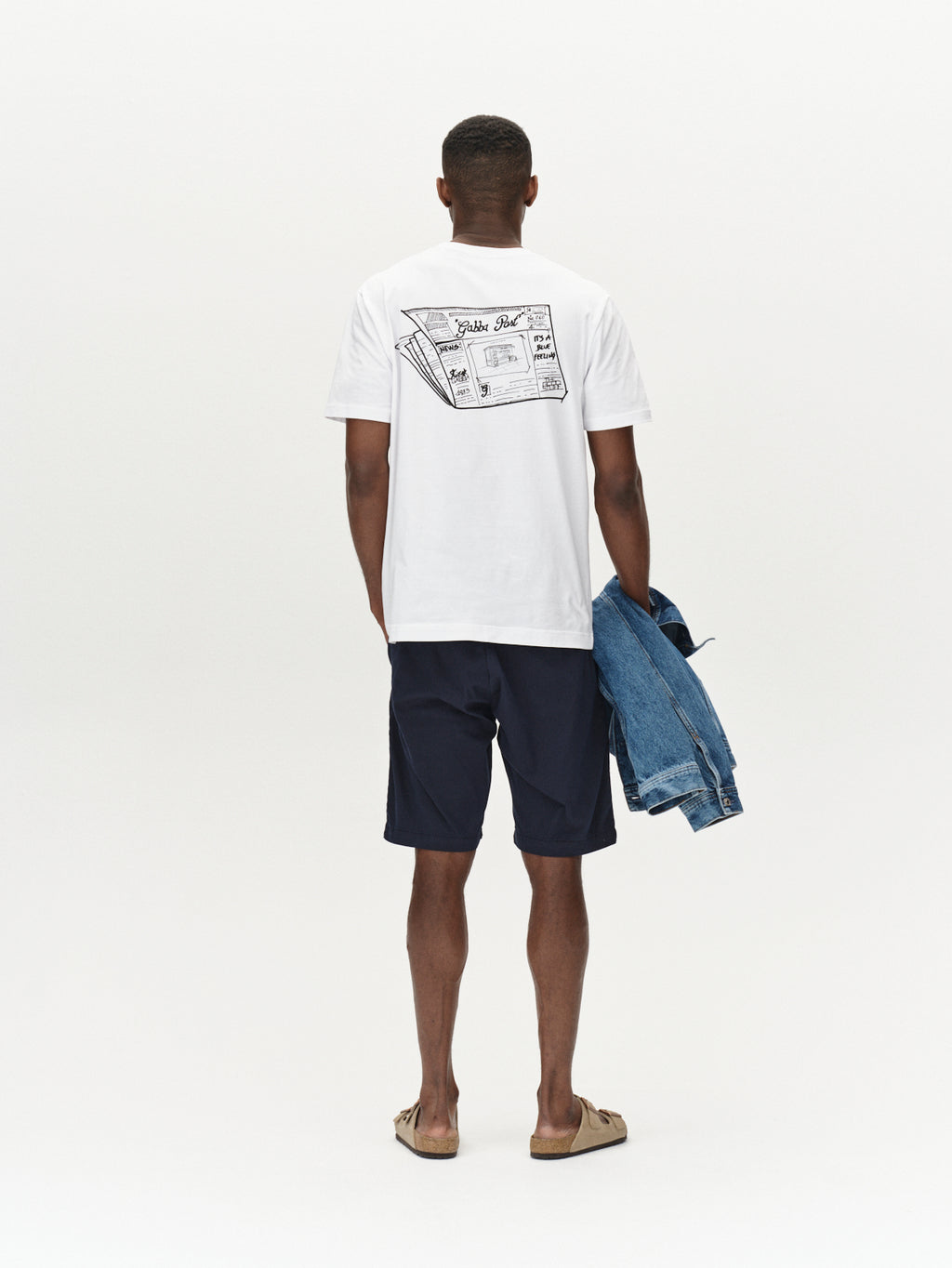 A person stands facing away, wearing a white t-shirt with a cassette tape graphic on the back, Gabbas Jet K3280 Dale Shorts in navy for all-day comfort, and sandals. They hold a denim jacket in their left hand against a plain white background.