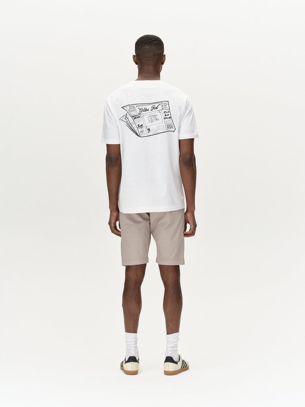 A man stands facing away, wearing a white T-shirt with a vintage newspaper graphic on the back. He pairs it with Gabbas Jet K3280 Dale Shorts in Grey Paloma, white socks, and beige sneakers with black stripes, all set against a plain white background for timeless appeal.